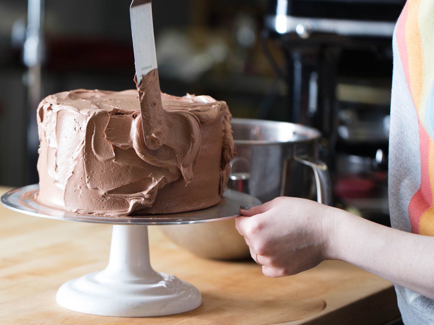 A person frosting a cake on a cakestand with chocolate Swiss buttercream.