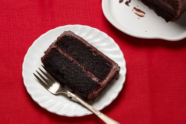 slice of cake on white plate, with cake to the side on white platter on a red fabric surface. 