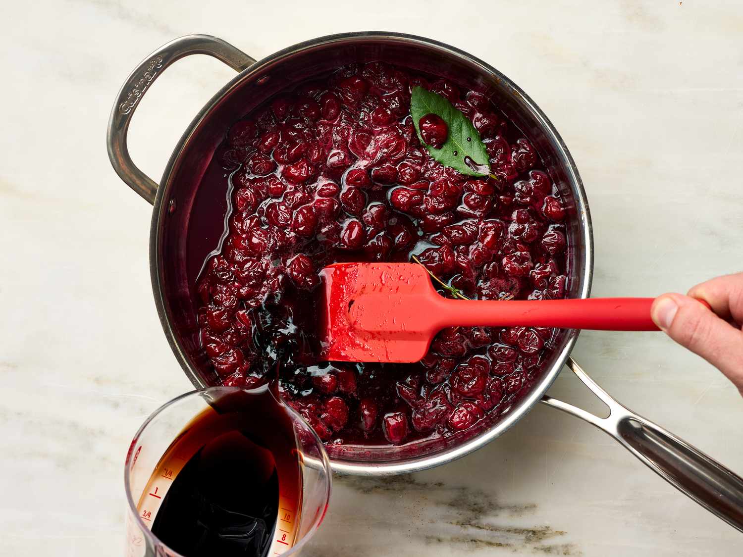 Simmering cherries being stirred in a saucepan with sauce being added from a measuring cup