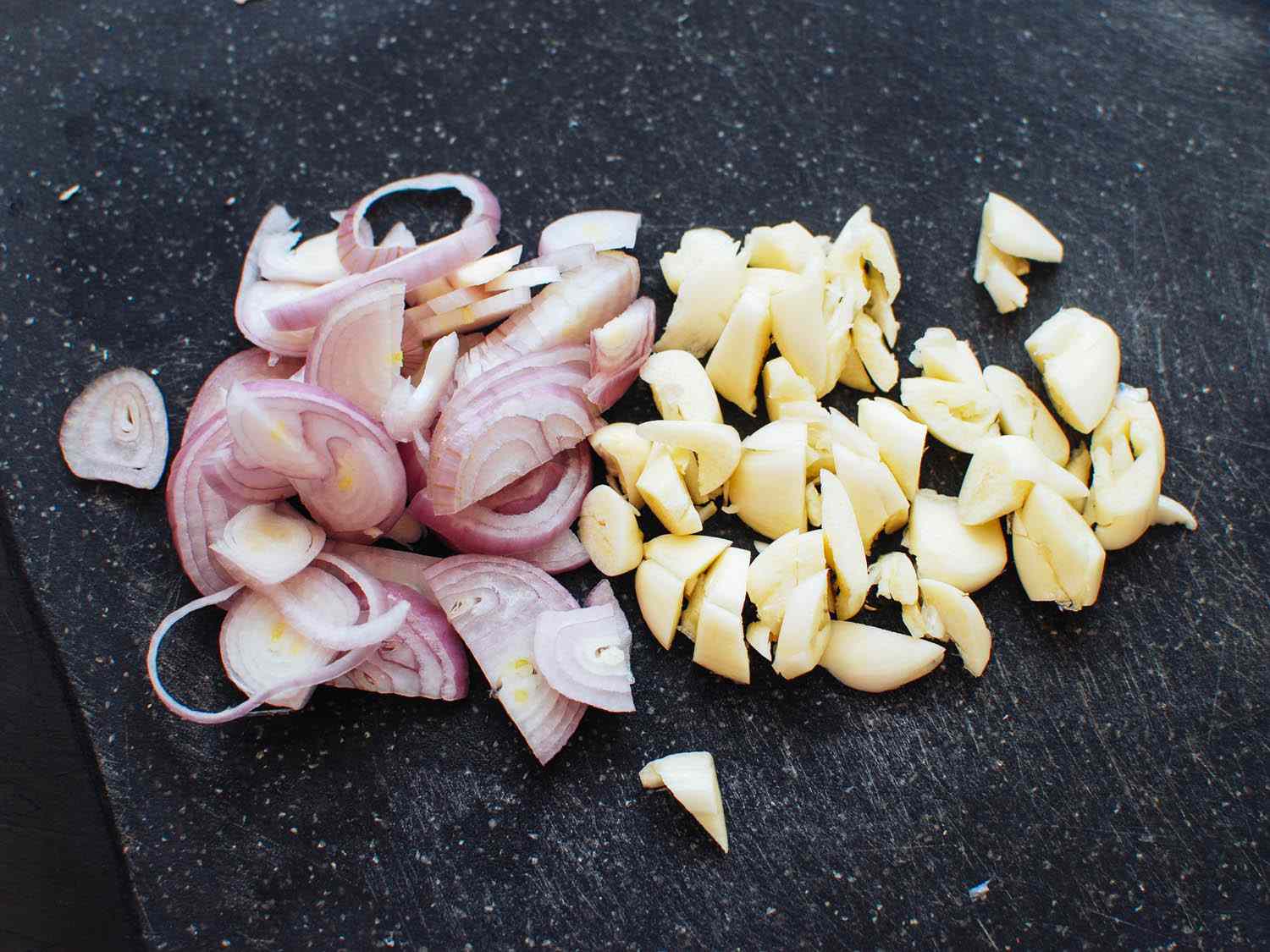Thinly sliced shallots and chopped garlic on a cutting board.