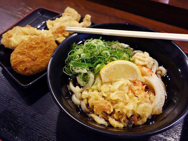 A bowl of udon noodles topped with scallions, lemon wedge, and battered tempura bits with assorted tempura on the side 