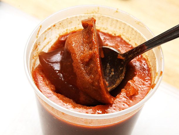 A quart-sized deli container filled with gelatinized stock. A spoon has broken through the layer of chile-tinted fat on top to reveal a dark brown broth.