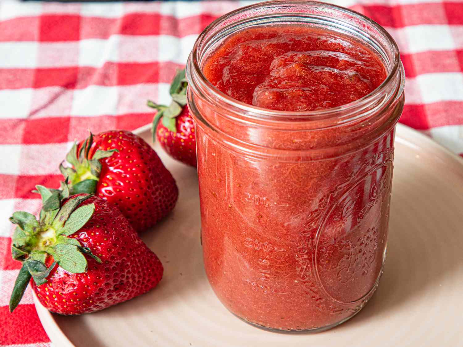 Glass jar of jam with whole strawberries on a plate and checkered tablecloth