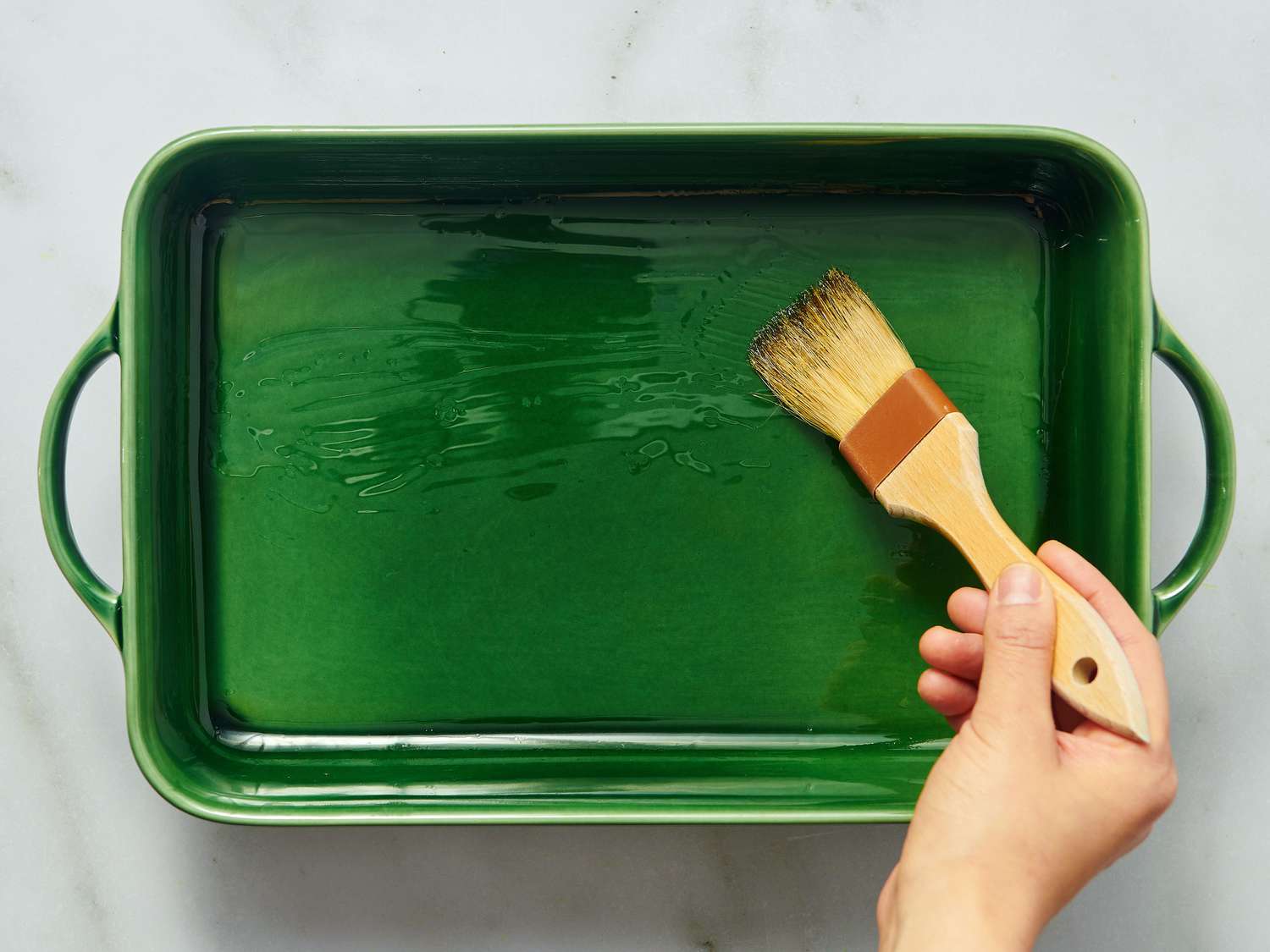 A hand brushing oil onto a green baking tray with a wooden brush