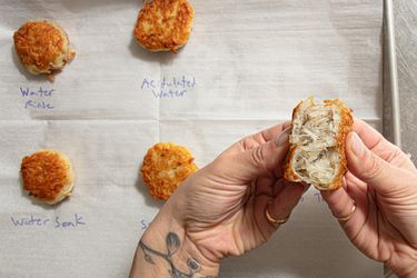 Hands holding a halved fried potato patty with several patties on a marked baking sheet in the background