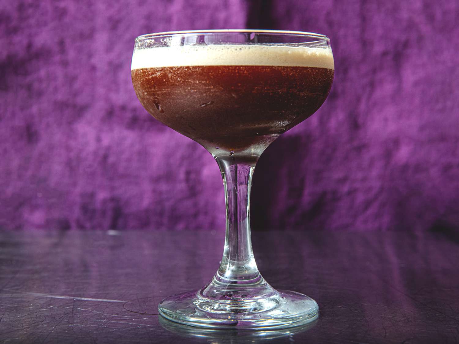 A glass with an espresso martini served on a tabletop against a purple background