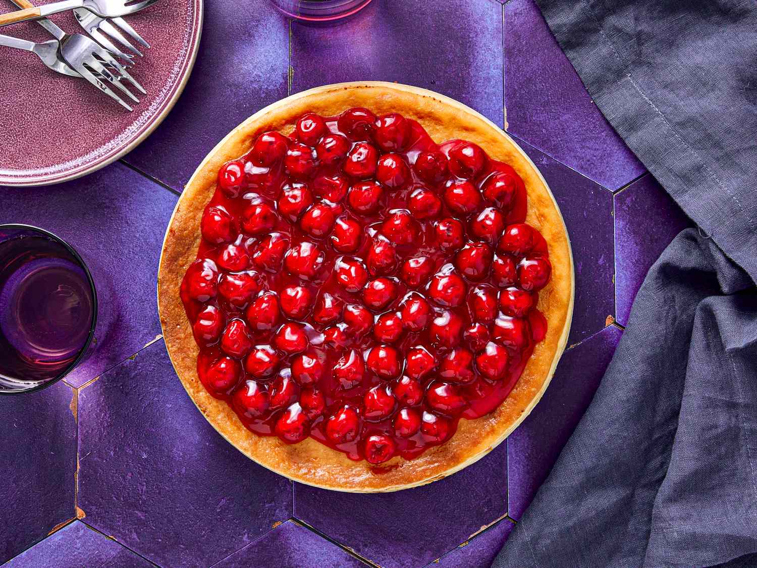 A cheesecake topped with cherries on a purple surface set with utensils and napkins for serving