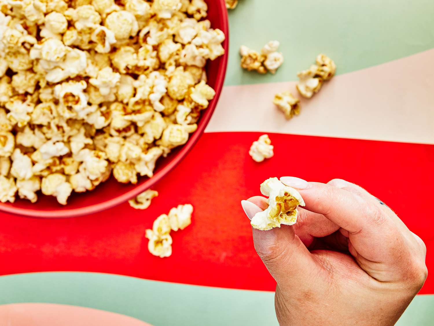 Red bowl full of Kettle corn on a red and green striped surface, with a hand in the foreground holding a popped kernel 
