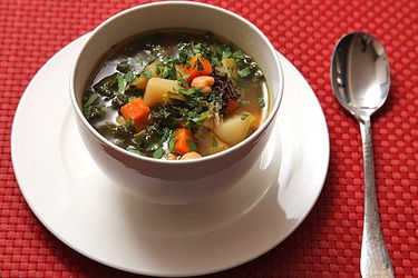 A bowl of hearty winter vegetable soup, served on a plate atop as red woven placemat.
