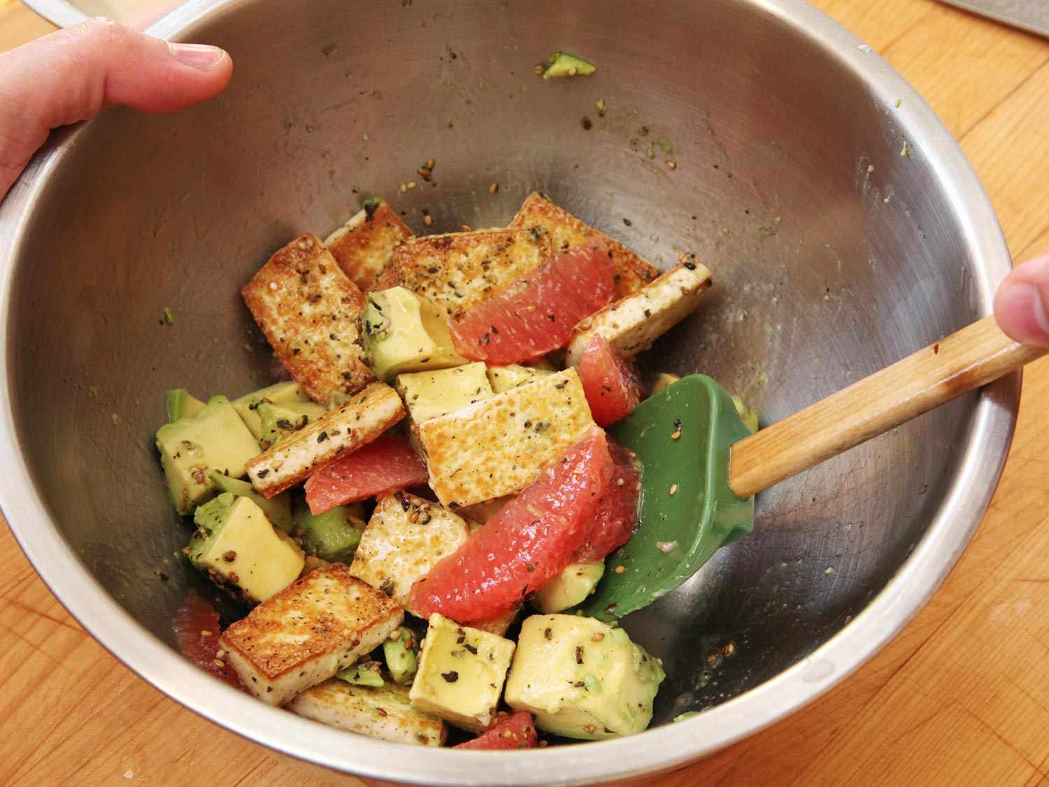 Tofu, avocado, and grapefruit being tossed with a silicone spatula in a metal mixing bowl.