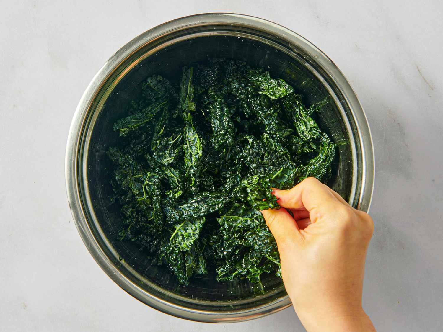 A hand massaging fresh kale leaves in a metal bowl