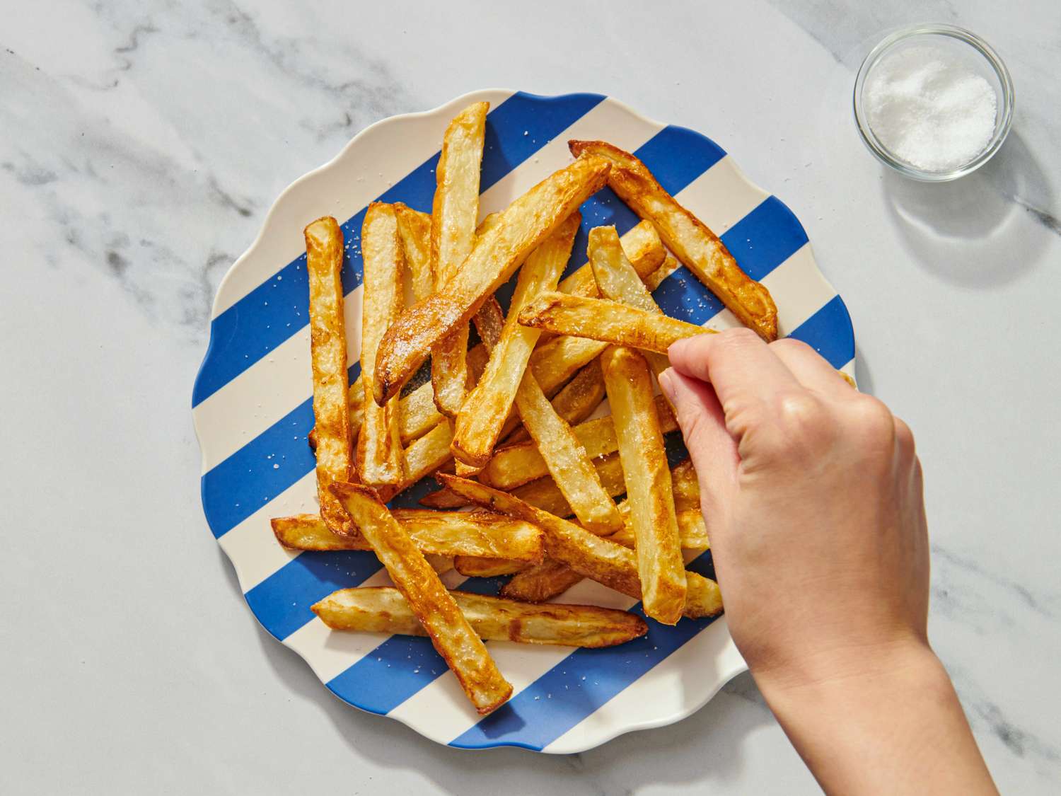A hand picking a French fry from a plate of cooked fries with a small bowl on the side