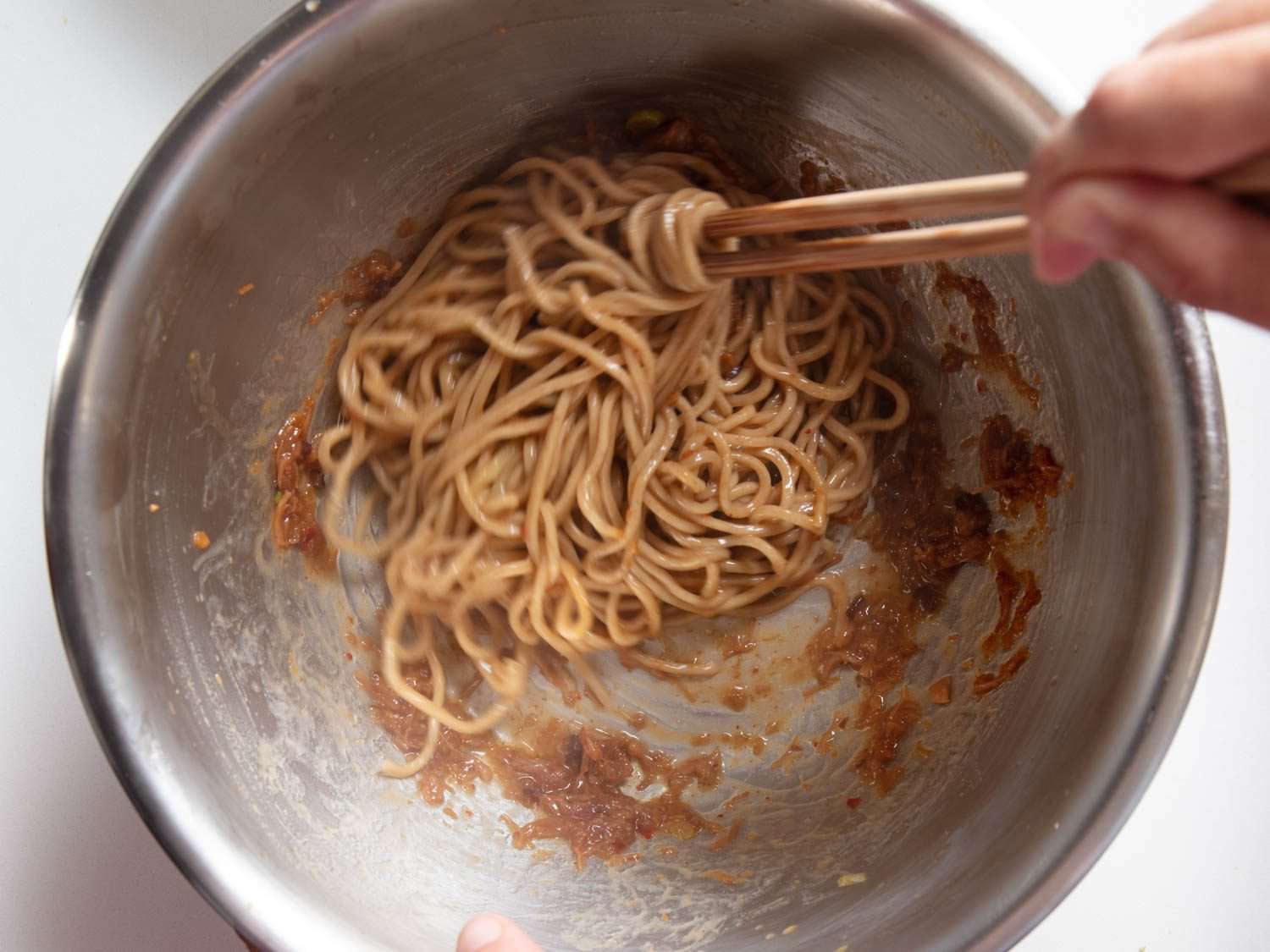 Noodles being tossed with XO sauce, soy sauce, rice vinegar, and pork fat in a mixing bowl.