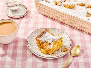 A piece of gooey butter cake on a plate with powdered sugar, with a cup of coffee and a serving tray of cake in the background