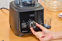 A close-up of a hand turning a nob on the Vitamix Ascent X2 resting on a wooden surface