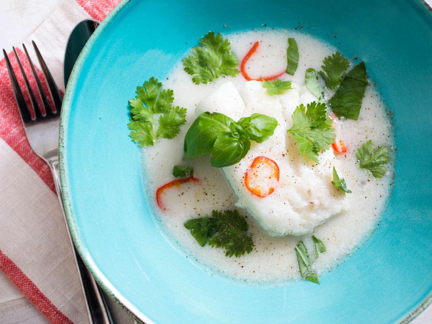 A bowl of poached cod in coconut milk with chiles, lime, and lemongrass garnished with fresh herbs.