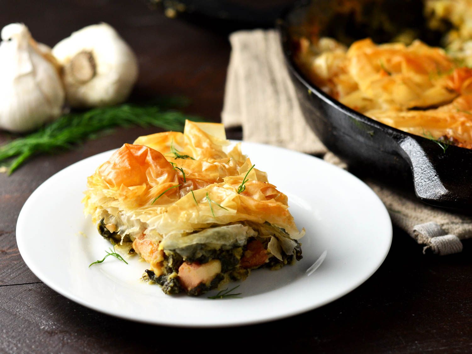 A square of chicken spanakopita on a white plate, with the full dish and two heads of garlic and fresh dill in the background.