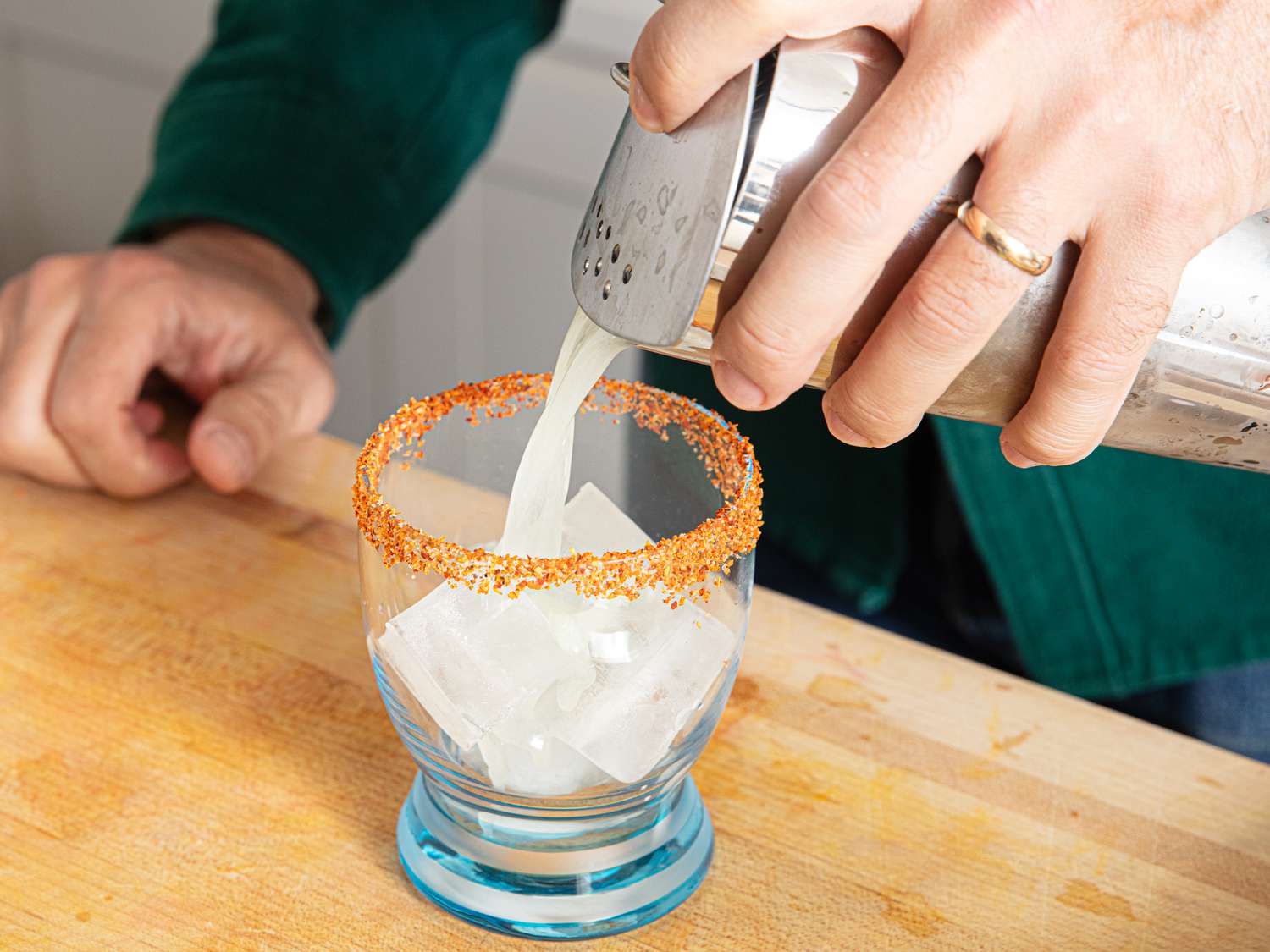 A person pouring a liquid from a shaker into a glass with ice cubes and a spiced rim