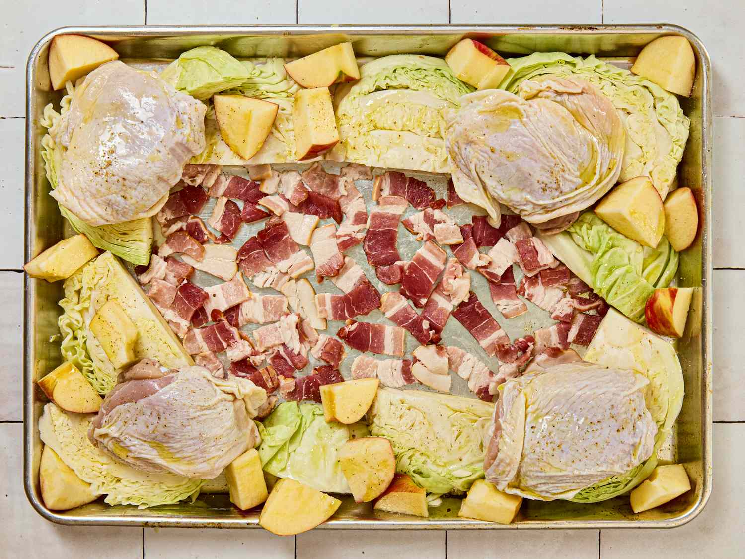 A sheet pan with chicken thighs cabbage wedges bacon pieces and apple slices arranged for roasting