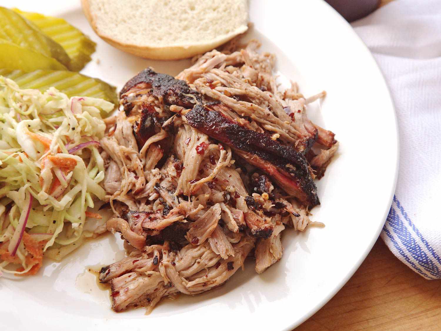 A plate of sous vide pulled pork with slaw, pickle slices, and a bun.