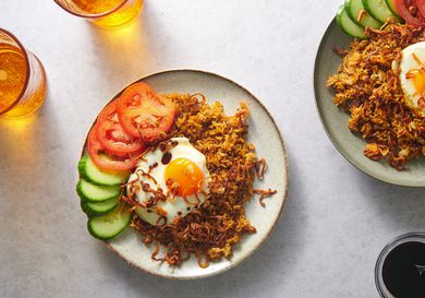 A plate of rice topped with a fried egg on a plate, garnished with cucumber and tomato slices, and showered with fried shallots. There is another plate to the right periphery of the image, as well as several glasses of liquid in the shot.