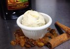 A scoop of rum raisin ice cream served in a ramekin. A bottle of dark rum, golden raisins, and cinnamon sticks are nearby.