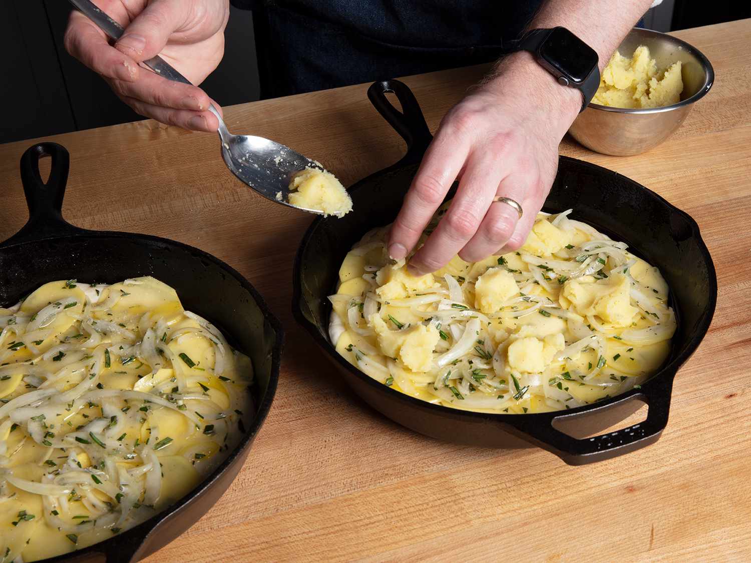 Two uncooked potato pizzas in side-by-side cast iron pans, with the rightmost pizza being topped with mashed potato by a pair of hands.