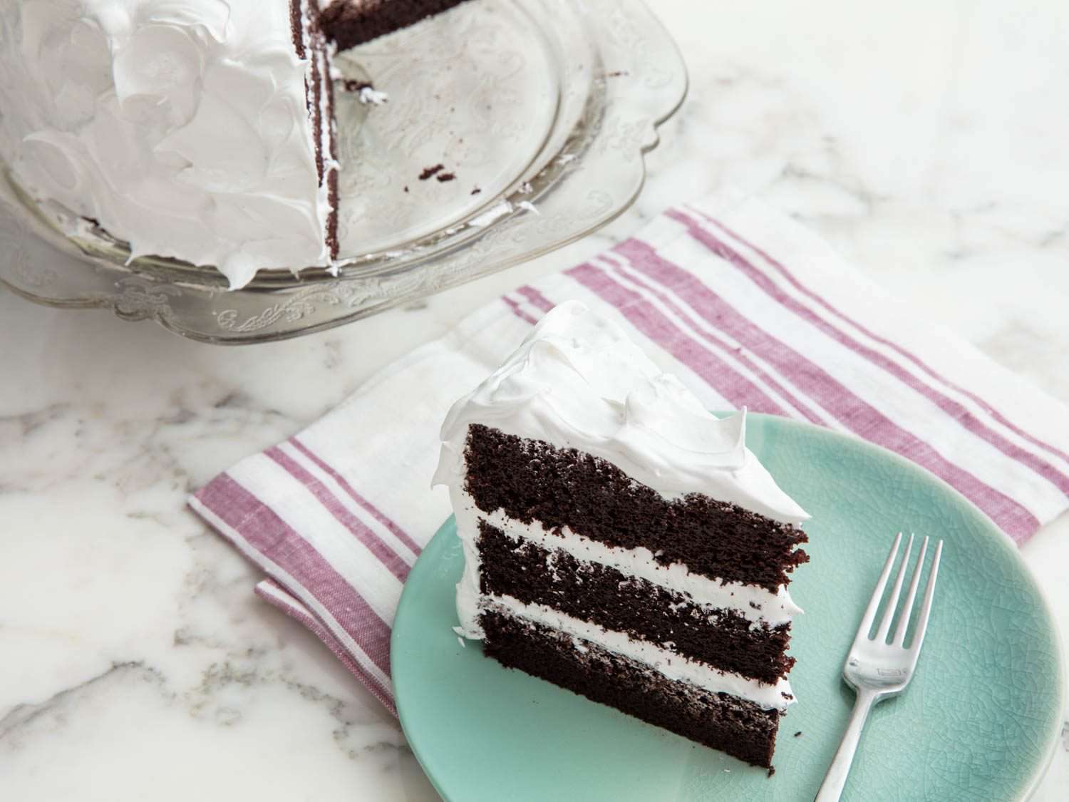 A three layer slice of devil's food cake, topped with fluffy white meringue, on a pale blue plate
