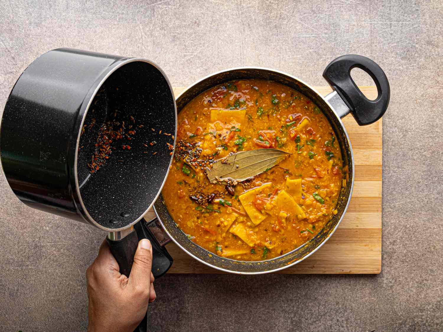 A person pouring spices into a pot of dal curry on a cutting board