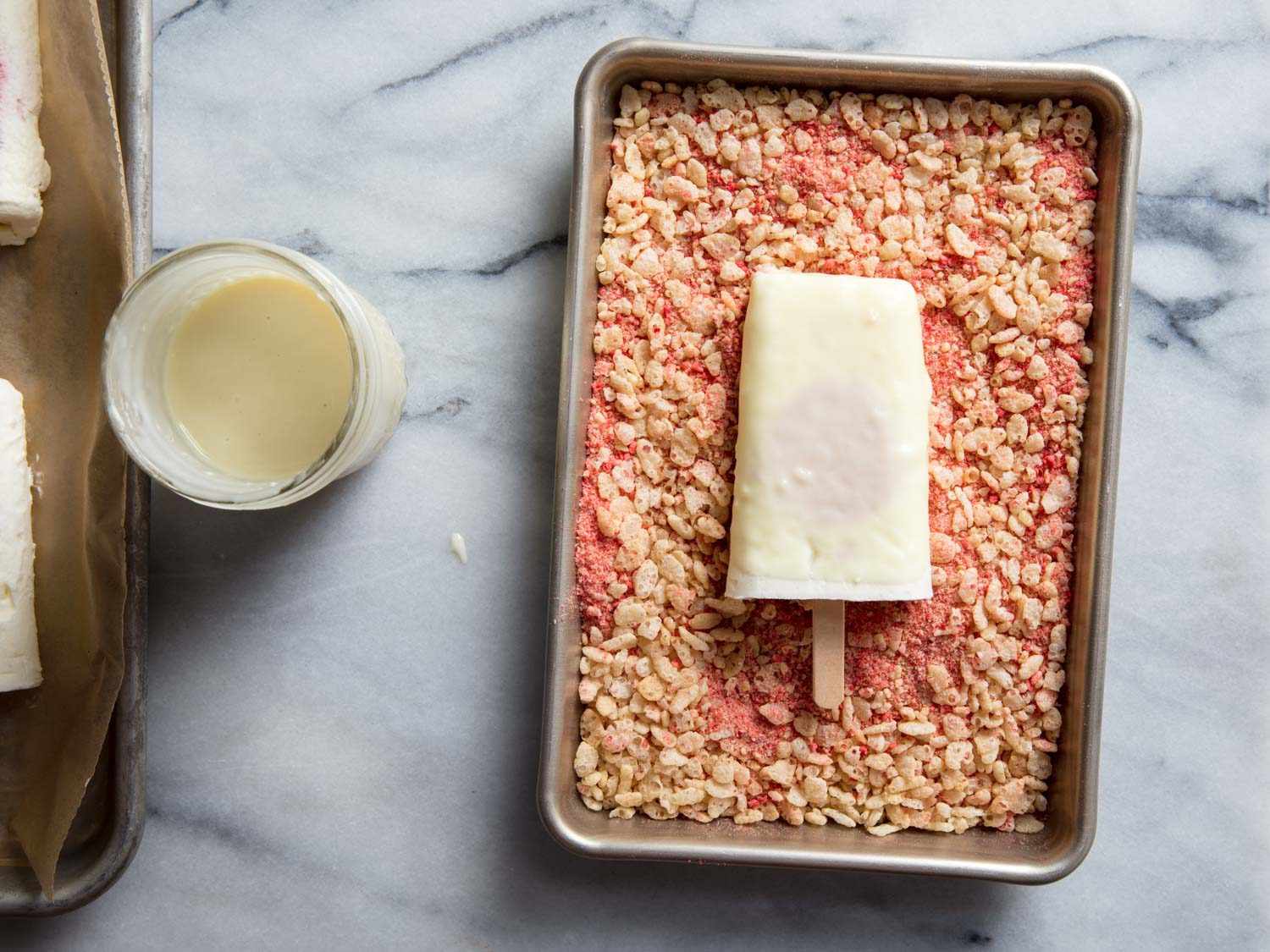 The dipped ice cream bar is on the crumb coating in a sheet pan, next to the glass containing the white chocolate coating. 