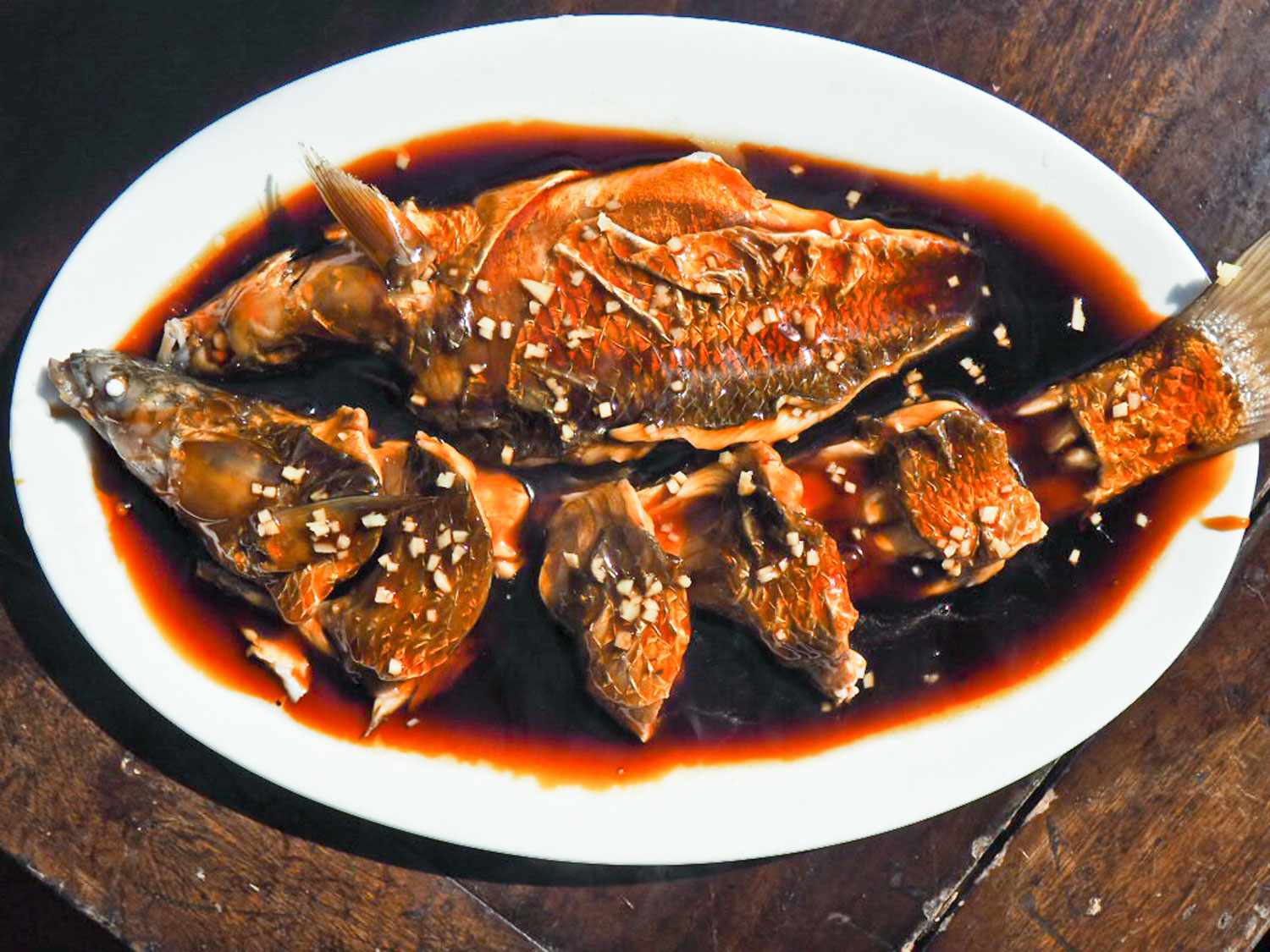 Overhead view of dayu darou—whole steamed fish—served on an oval platter ands smothered in a dark brown sauce tinted red from a thin layer of chile oil on the surface.