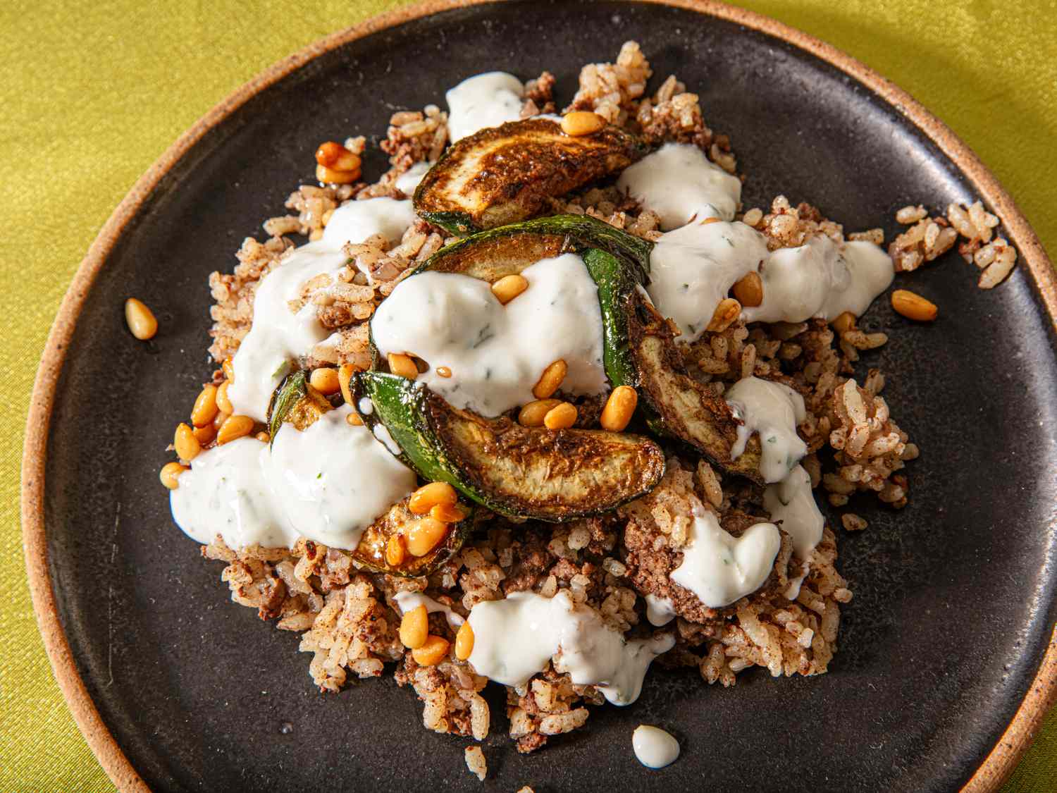 A plate of rice topped with roasted zucchini slices pine nuts and creamy sauce served on a dark dish