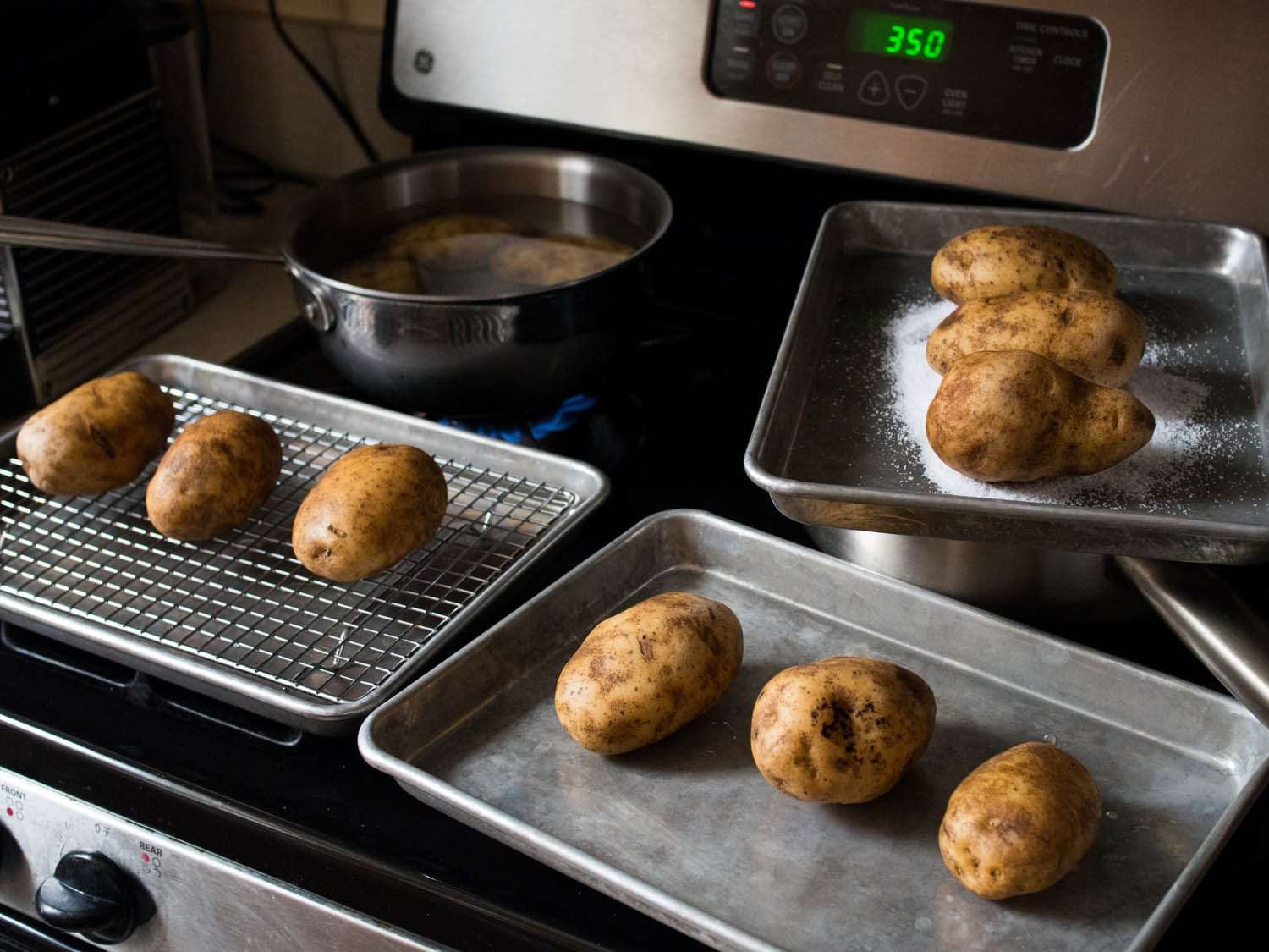 Three pans with three potatoes each: one has a rack, one is just the pan, and the third the potatoes sit on a pile of salt. There's also a pot filled with water and three potatoes. 