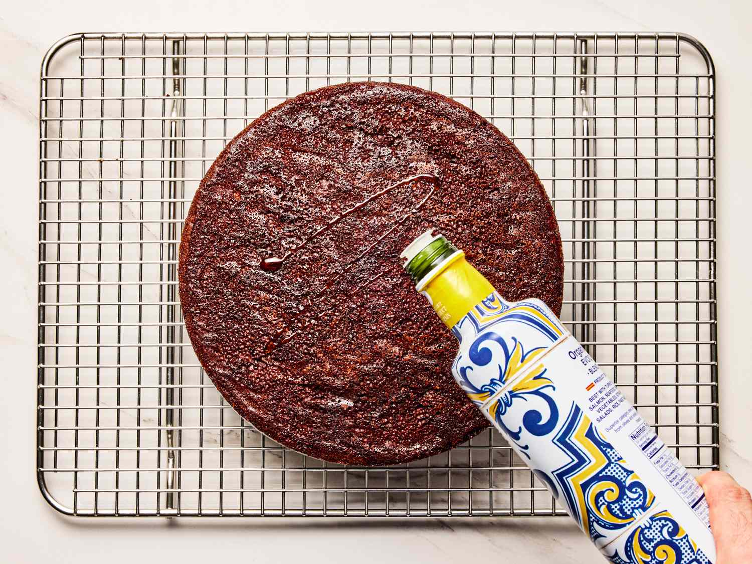 A chocolate cake on a cooling rack being drizzled with olive oil from a decorative bottle