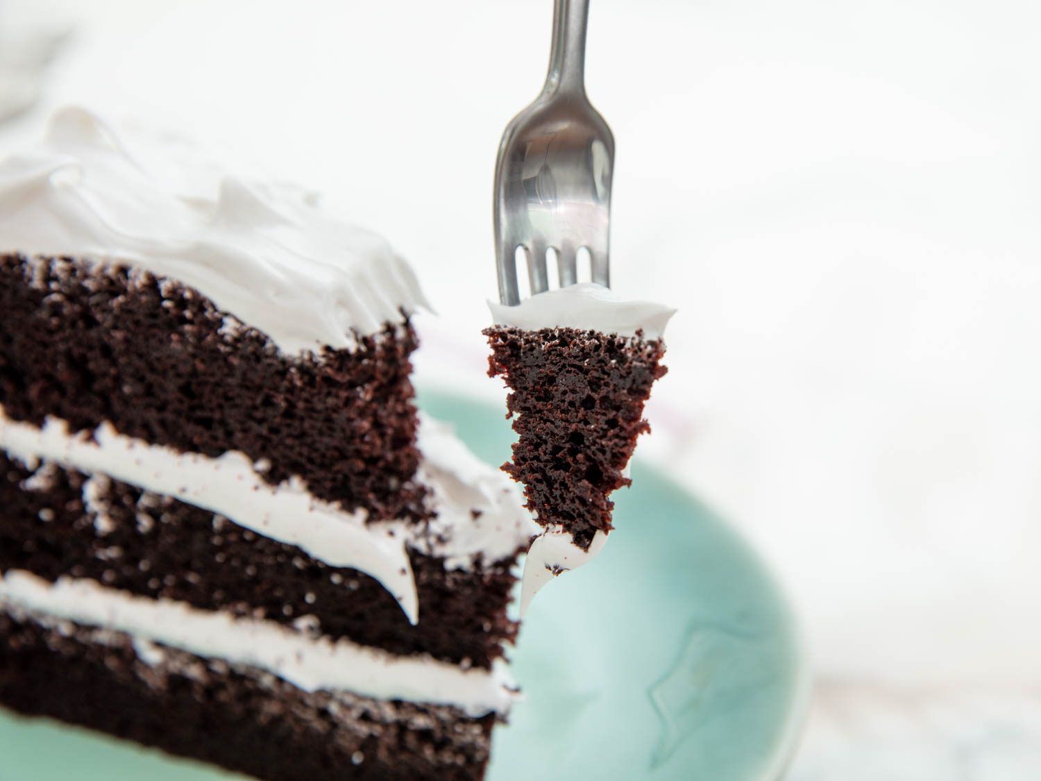 A three layer slice of chocolate cake frosted with a fluffy white meringue, with a single bite skewered by a fork