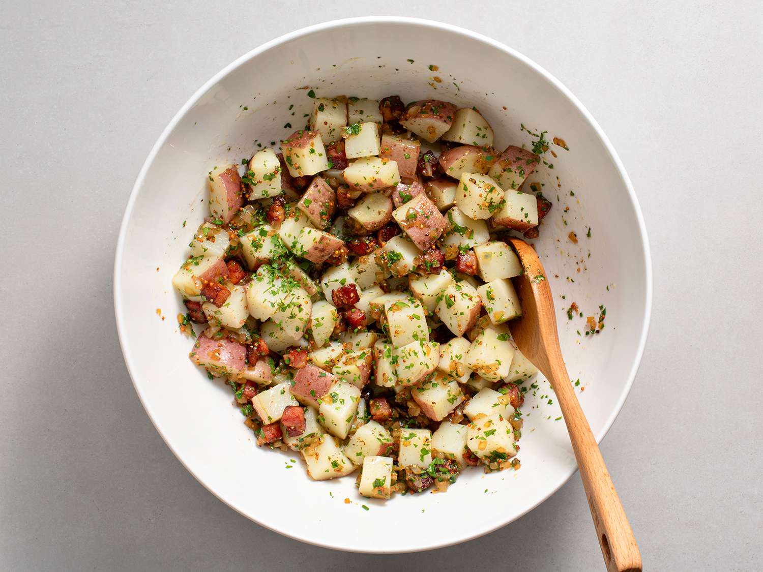 Cooked potatoes being tossed with vinaigrette in a white ceramic bowl, with a wooden spoon.