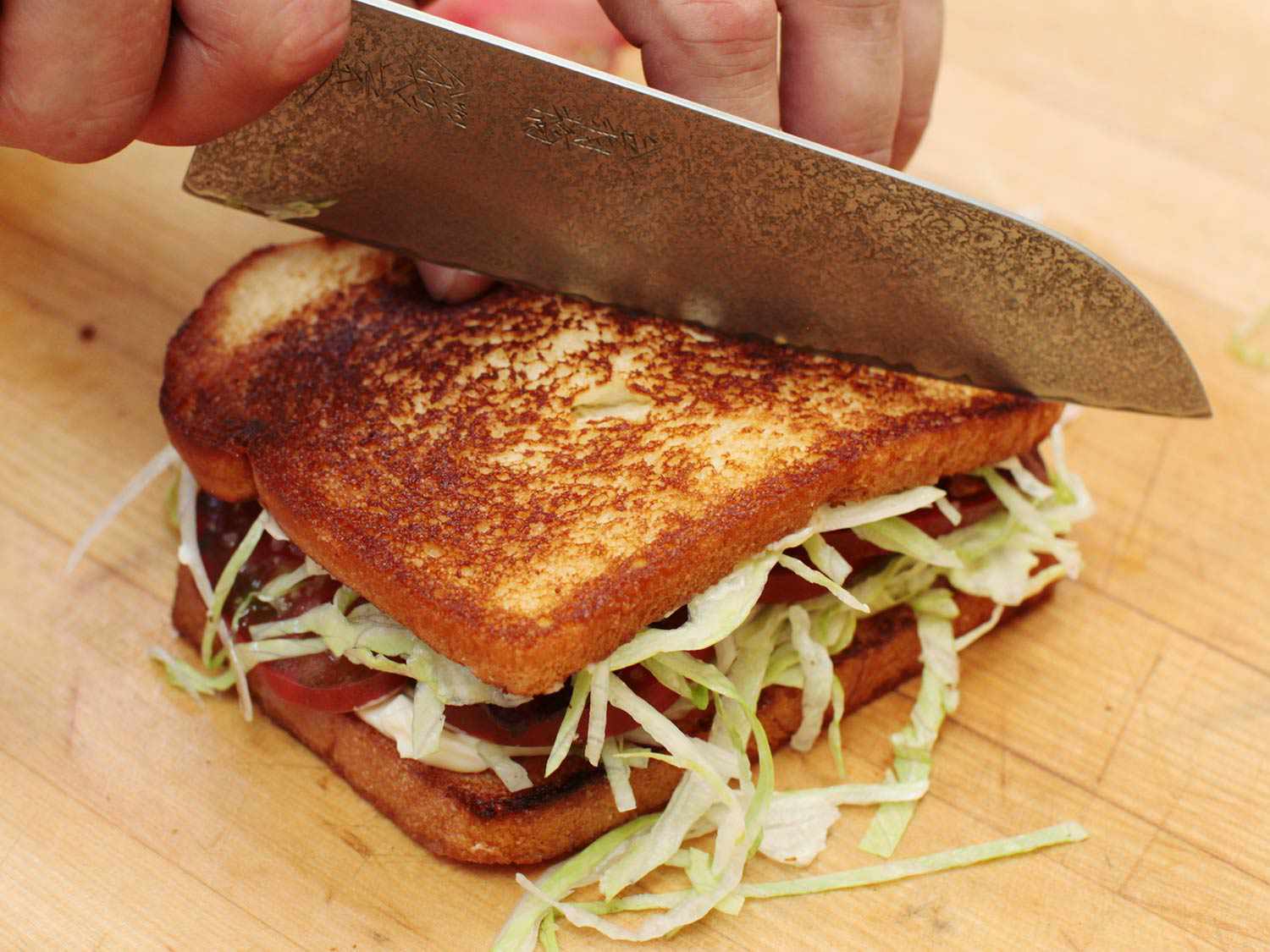A chef's knife slicing a finished BLT sandwich in half.