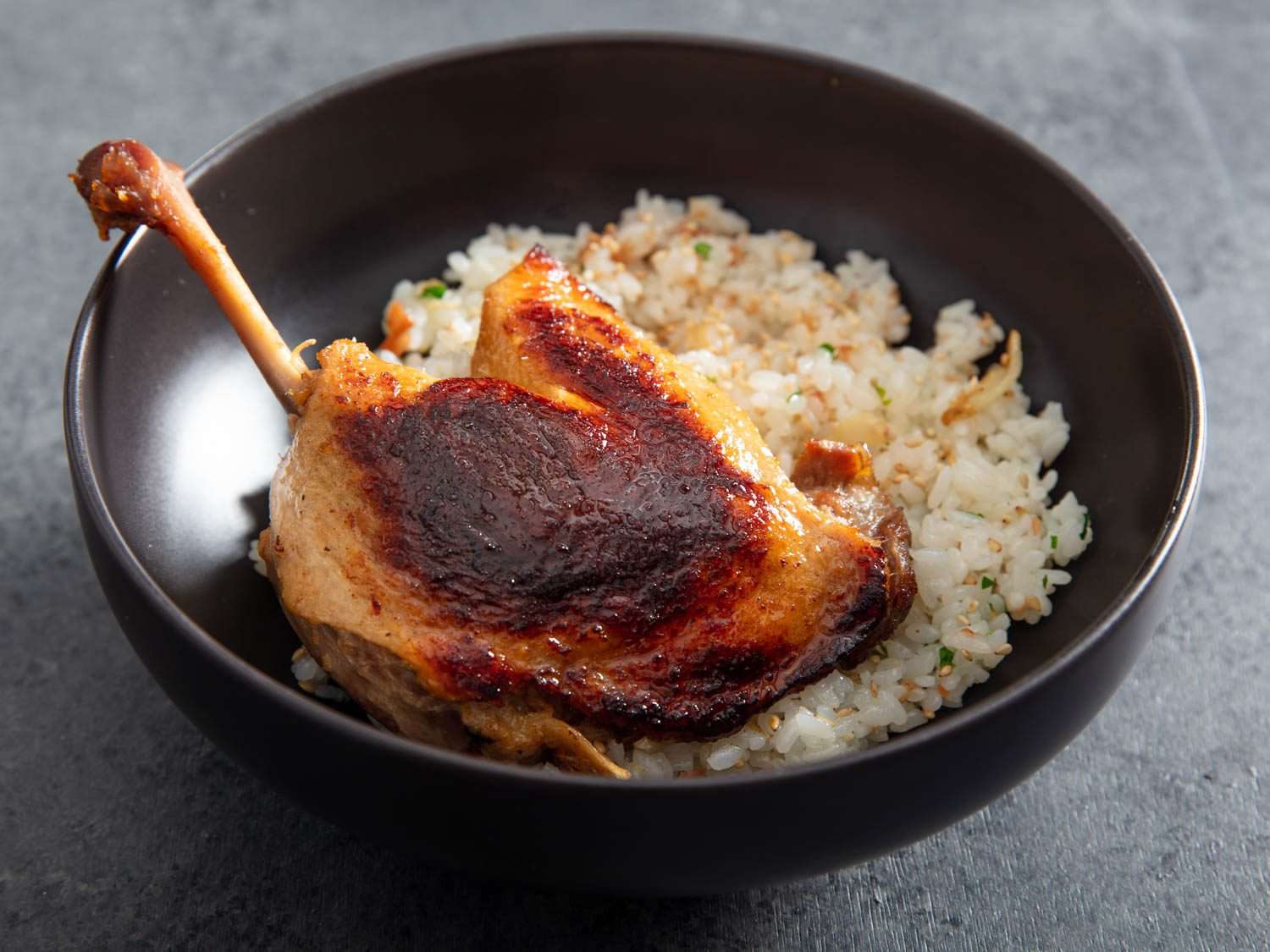 Closeup side view of a crisped koji confit duck leg in a bowl with rice.