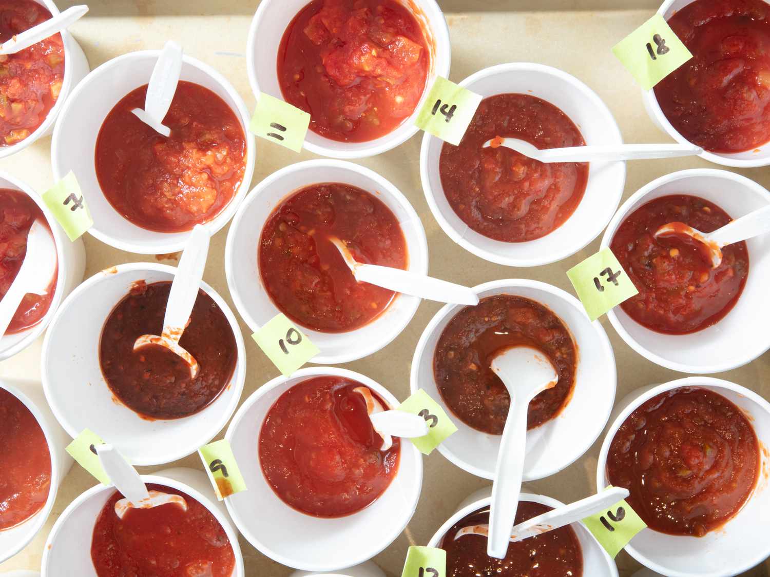 Multiple labeled cups of salsa with spoons for a taste test