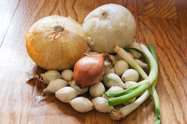 A pile of different types of whole, unpeeled onions on a cutting board. 