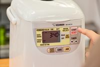 A close-up of a finger pressing a button on the Zojirushi Home Bakery Mini Breadmaker