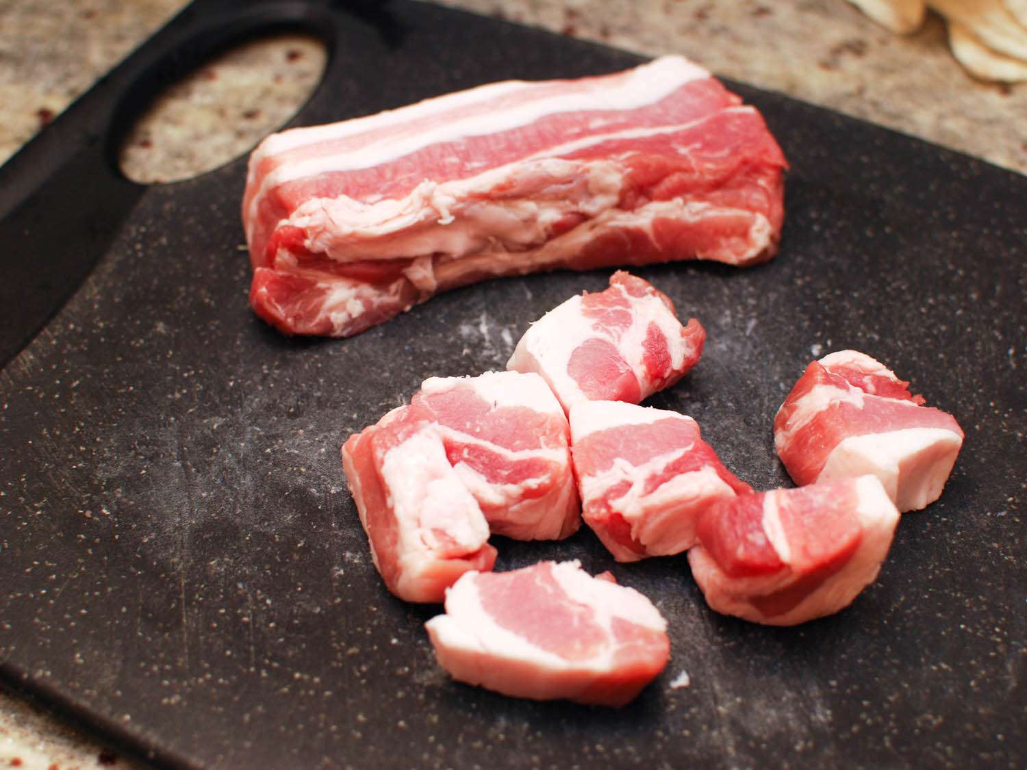 A thick slab of pork belly is sliced into chunks on a cutting board.