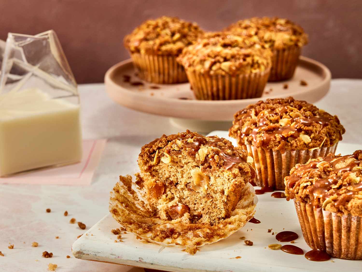 apple pie muffins on a white tray, one with a bite taken out of it, with 3 muffins on a circular tray in the background. A clear glass of milk to the side on pink napkins. 