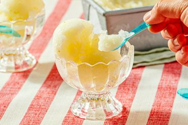 A hand scooping lemon sorbet from a glass bowl with a blue spoon placed on a red and white striped tablecloth