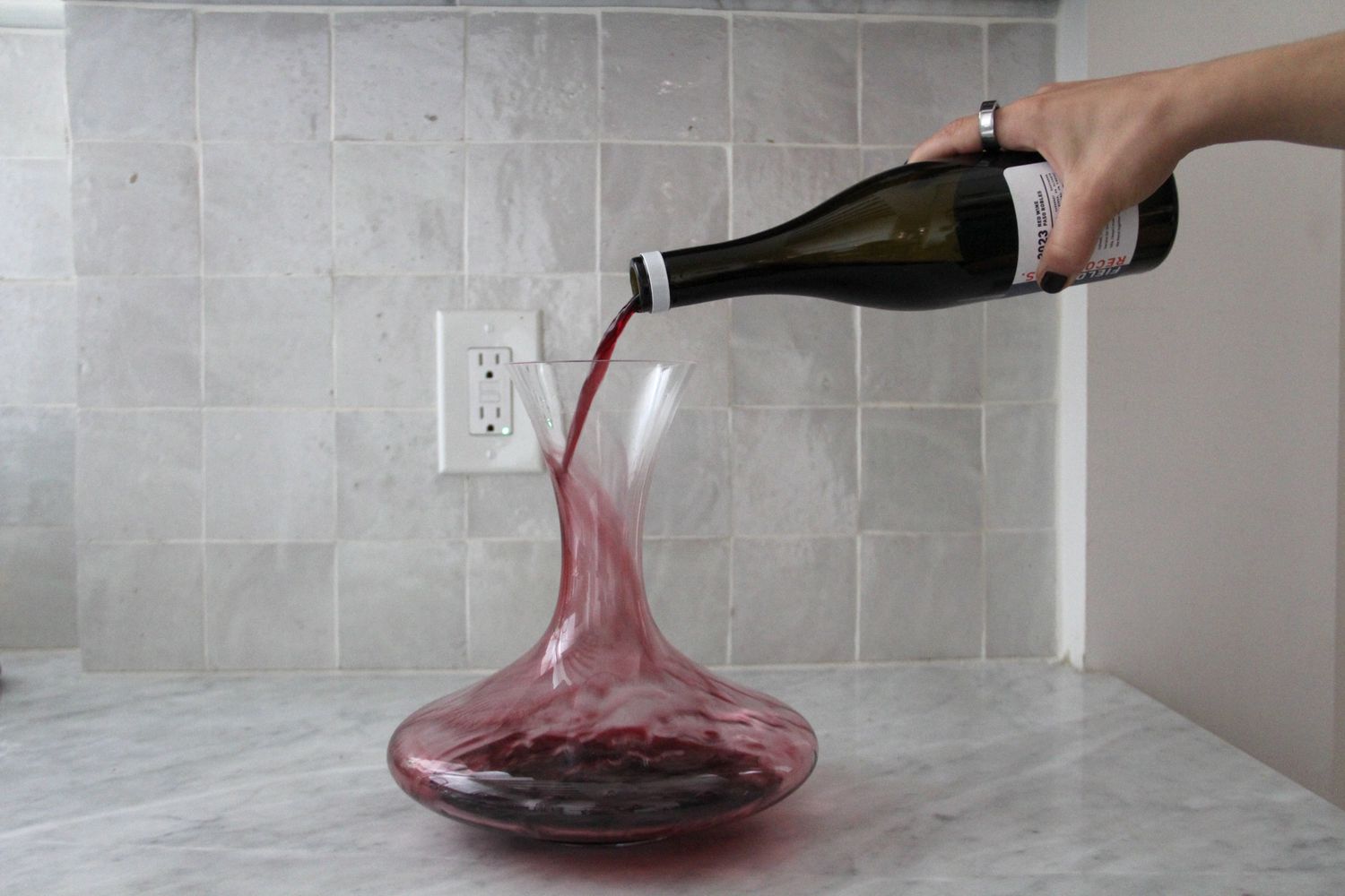 A bottle of red wine being poured into a glass wine decanter on a marble counter