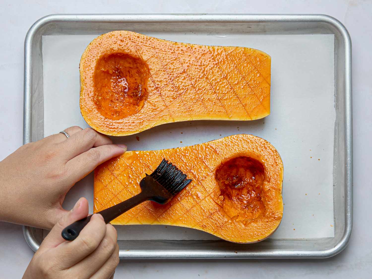 Hand using a brush to apply a seasoning glaze to halved butternut squash on a baking tray