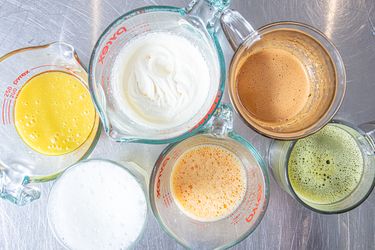Six different beverages including milky and frothy textures displayed in measuring cups on a metal surface