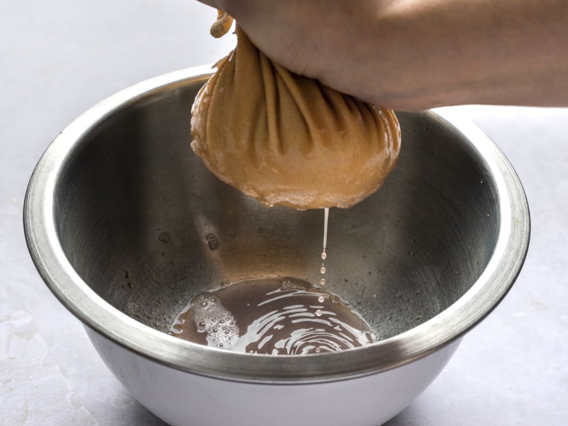 Shredded potatoes being squeezed in a dish towel, with excess liquid running off into a metal bowl.