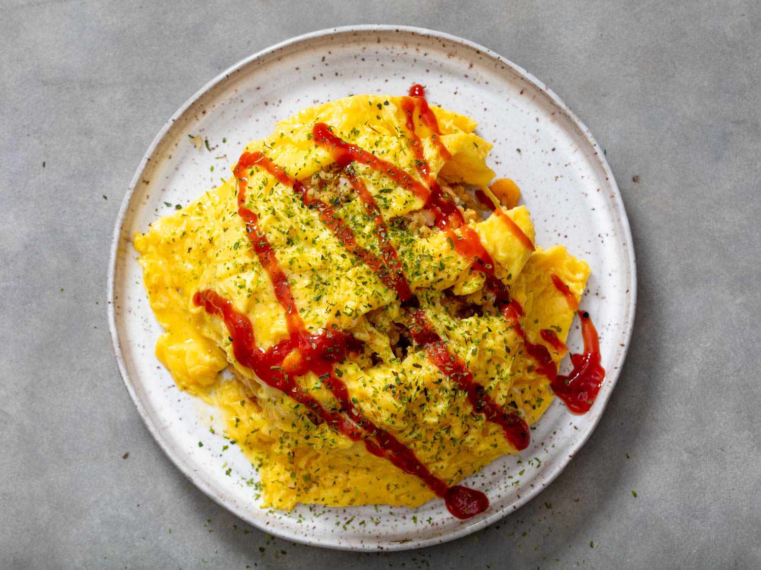 An unfolded omelet has been blanketed over the fried rice and topped with a squiggle of ketchup and furikake seasoning.