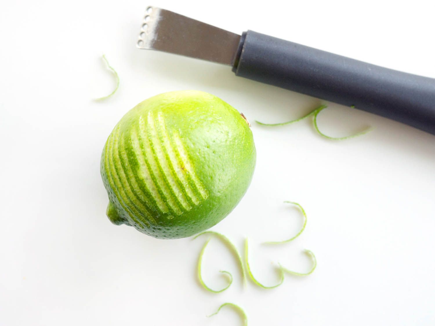 Closeup of a lime that has been partially zested. A zester and shreds of zest are scattered on the white surface below.